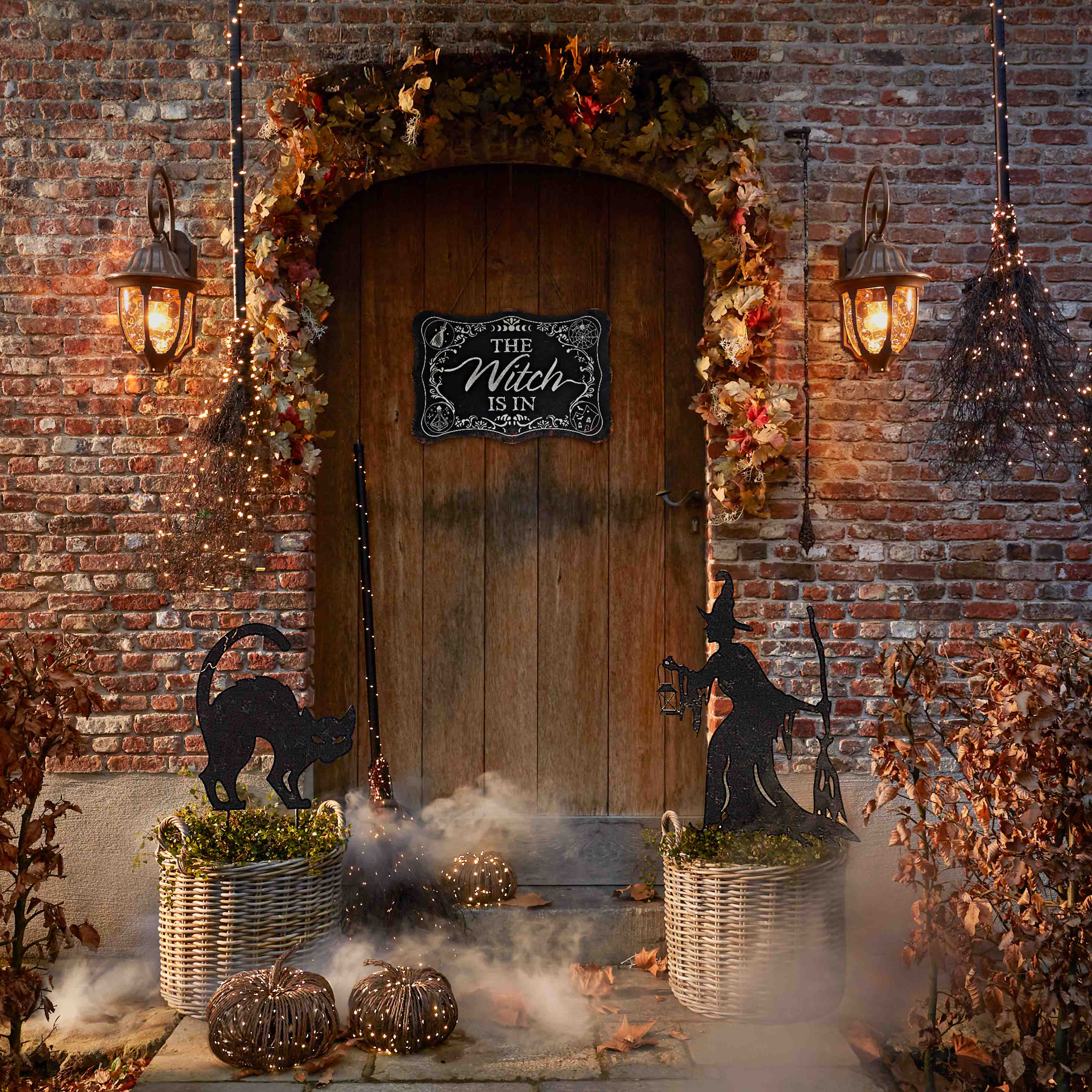 Decorazione di Halloween con gatto nero, strega e lanterne davanti a porta in legno e muro di mattoni