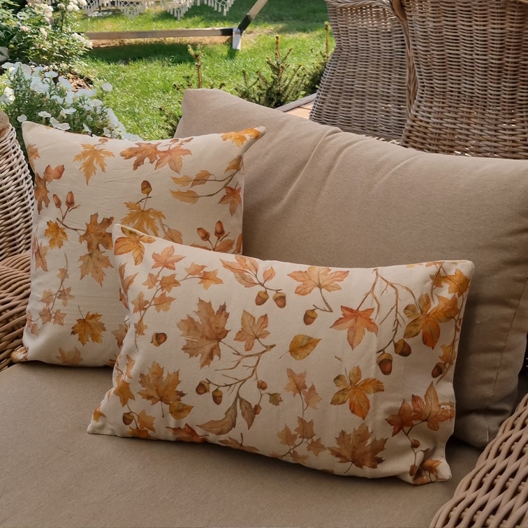 Cuscini decorativi in cotone beige con stampa di foglie autunnali e ghiande su divano da giardino in rattan