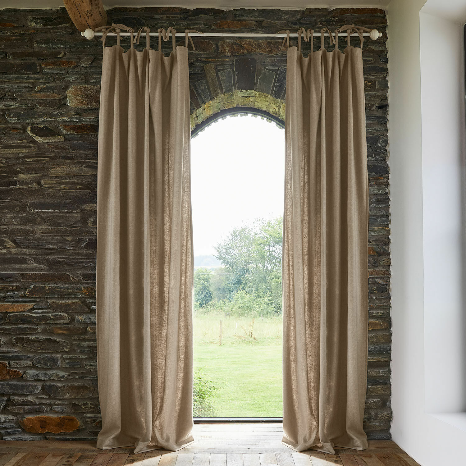 Tenda Seleneia Tende beige in tessuto naturale su bastone in interno in pietra con vista sul giardino