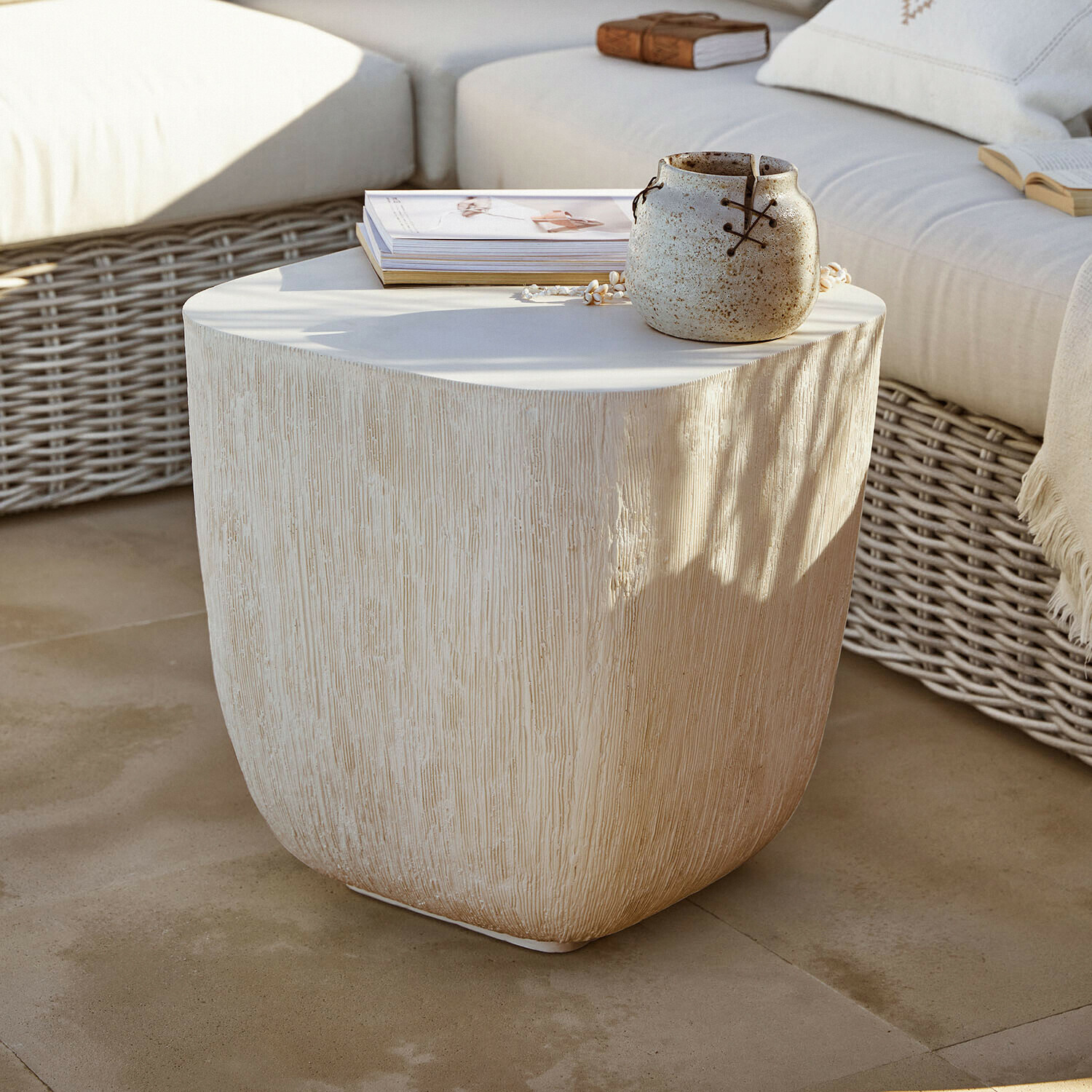 Tavolino beige con texture legno, vaso in ceramica e libri su terrazza con divano in rattan