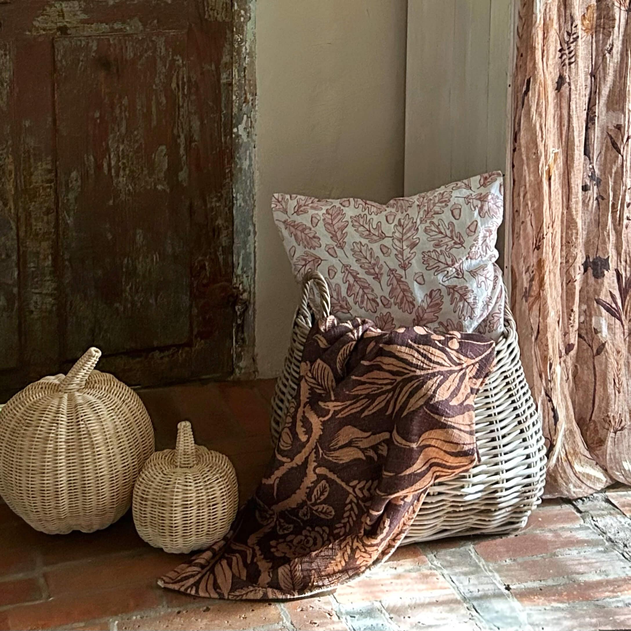 Cesto in vimini bianco con cuscino a foglie e coperta marrone con motivi floreali su pavimento in mattoni