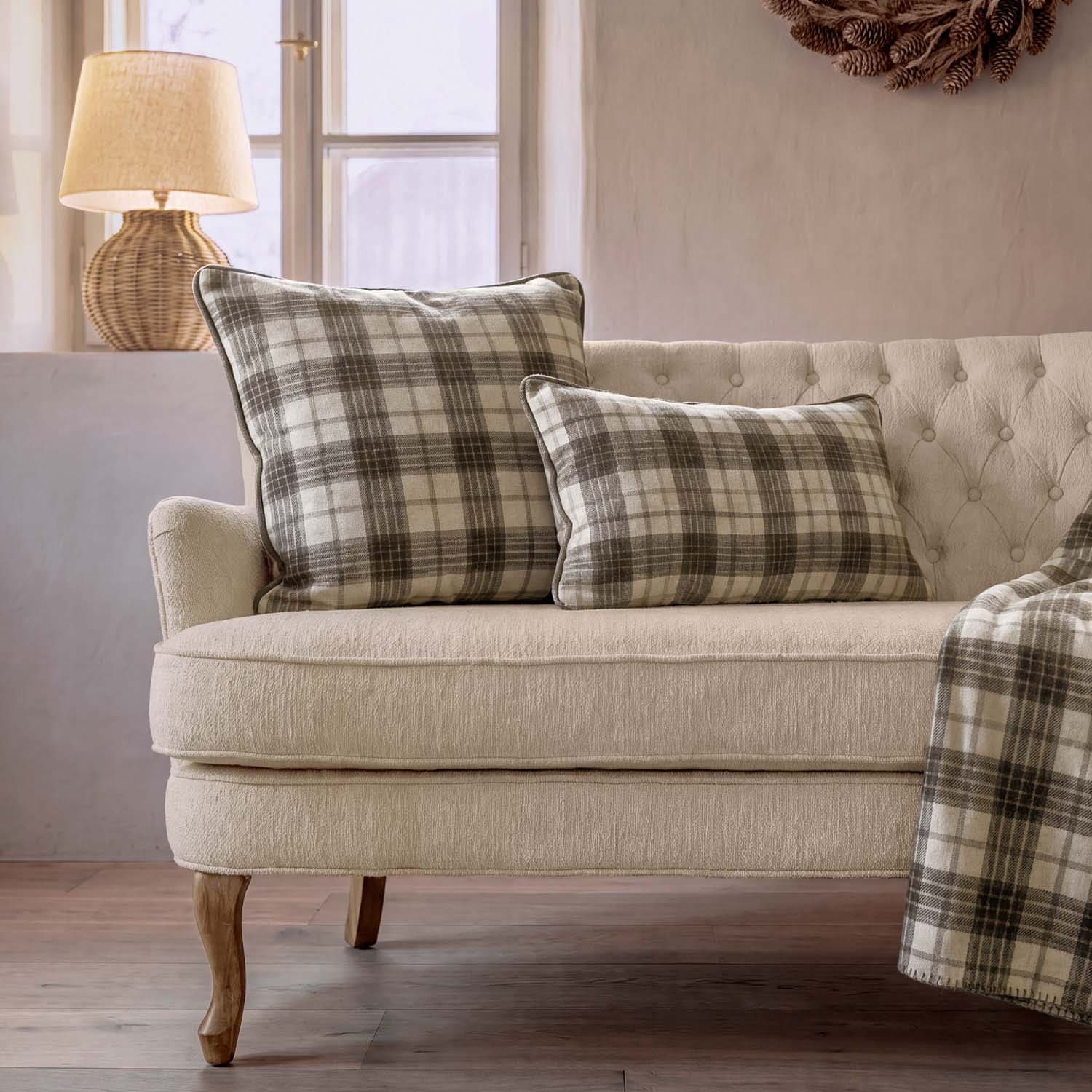 Divano beige con due cuscini a quadri grigi e crema, lampada in vimini vicino alla finestra
