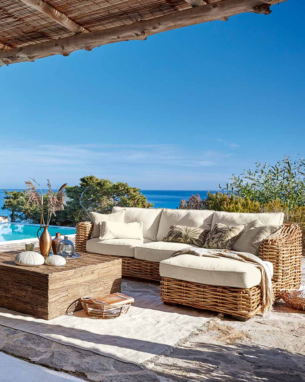 Set di mobili da giardino in rattan con cuscini crema e tavolo in legno su terrazza con vista mare