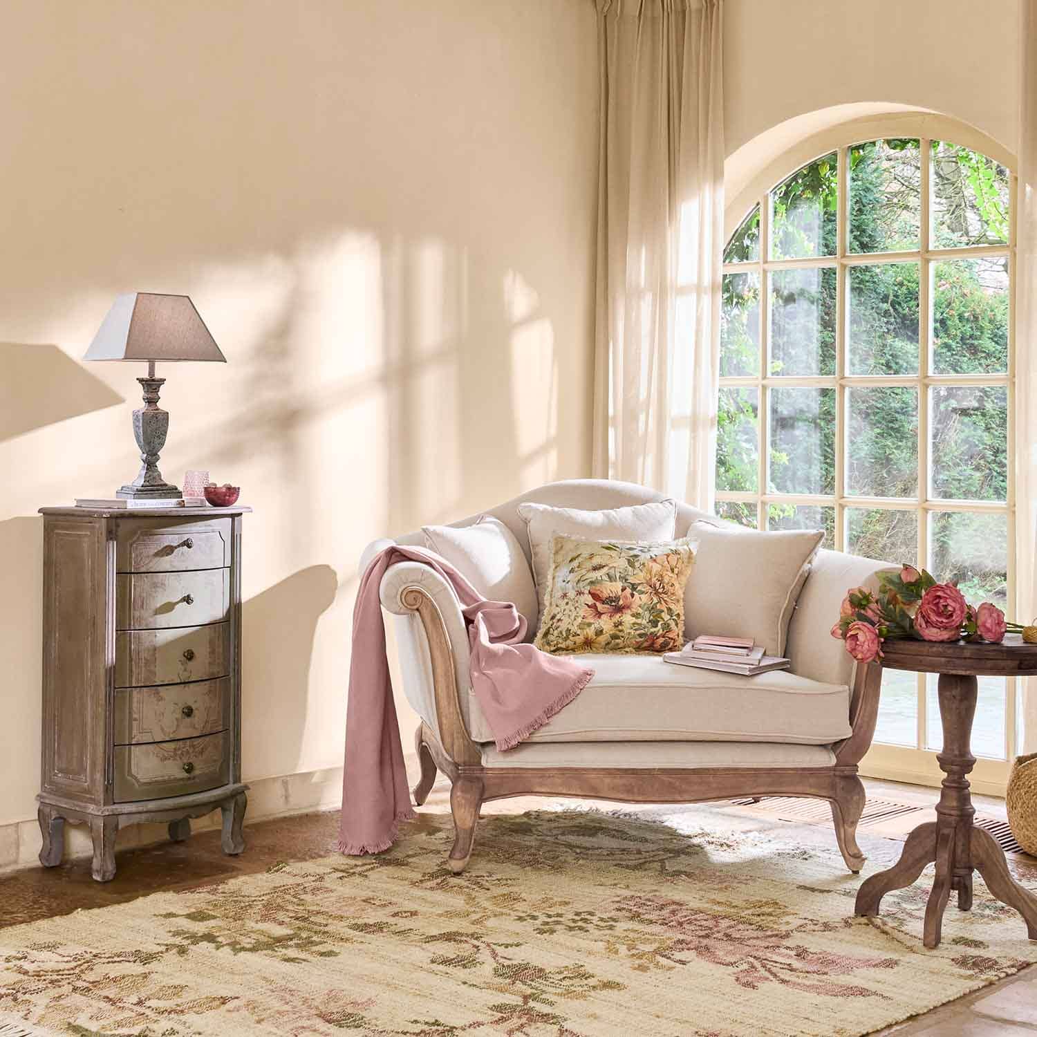 Divano crema con struttura in legno, cuscino floreale e coperta rosa in soggiorno luminoso con grande finestra