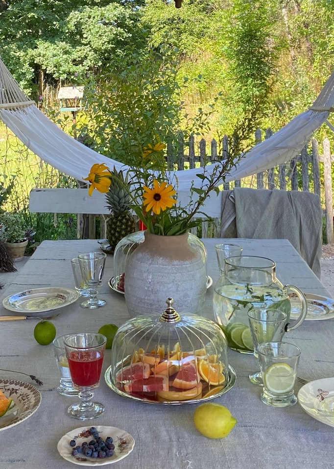 Tavolo all'aperto con tovaglia grigia, vaso con fiori gialli e campana di vetro con frutta, atmosfera rilassante in giardino