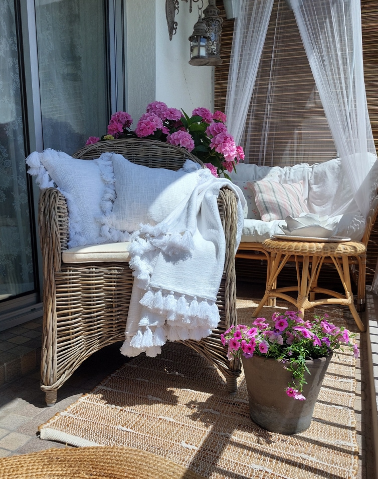 Poltrona in rattan con coperta bianca e cuscini, fiori rosa e tavolo in materiali naturali sulla terrazza