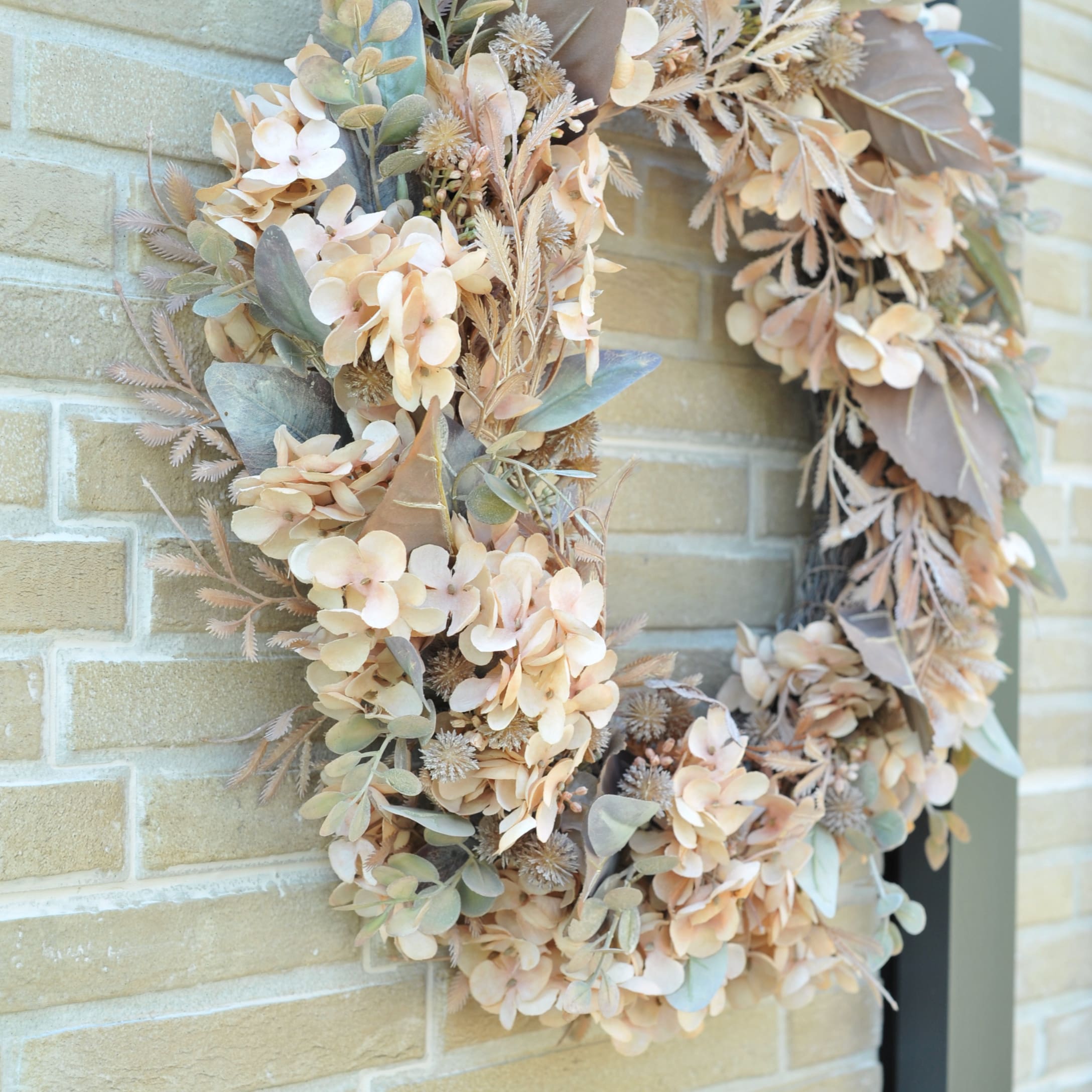Corona decorativa autunnale con foglie artificiali beige e verdi su muro di mattoni