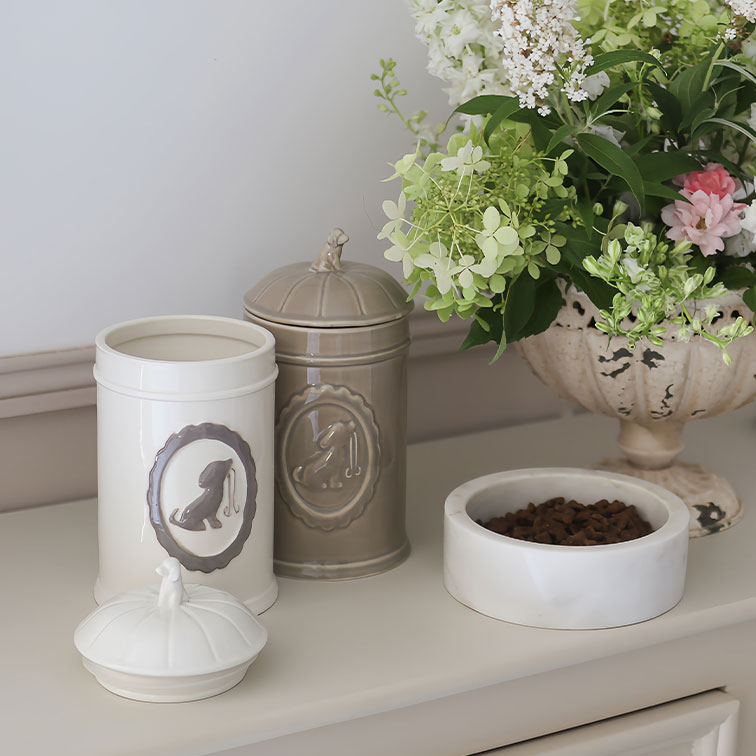 Contenitori in ceramica crema e beige per cibo cane accanto a ciotola bianca con crocchette e fiori