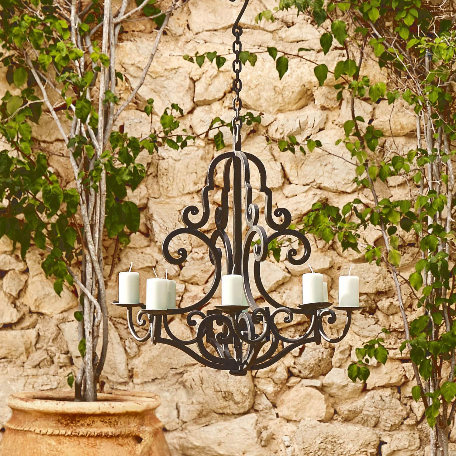 Lampadario per candele Geneuve Lampadario in metallo nero con sei candele bianche davanti a muro di pietra e foglie verdi