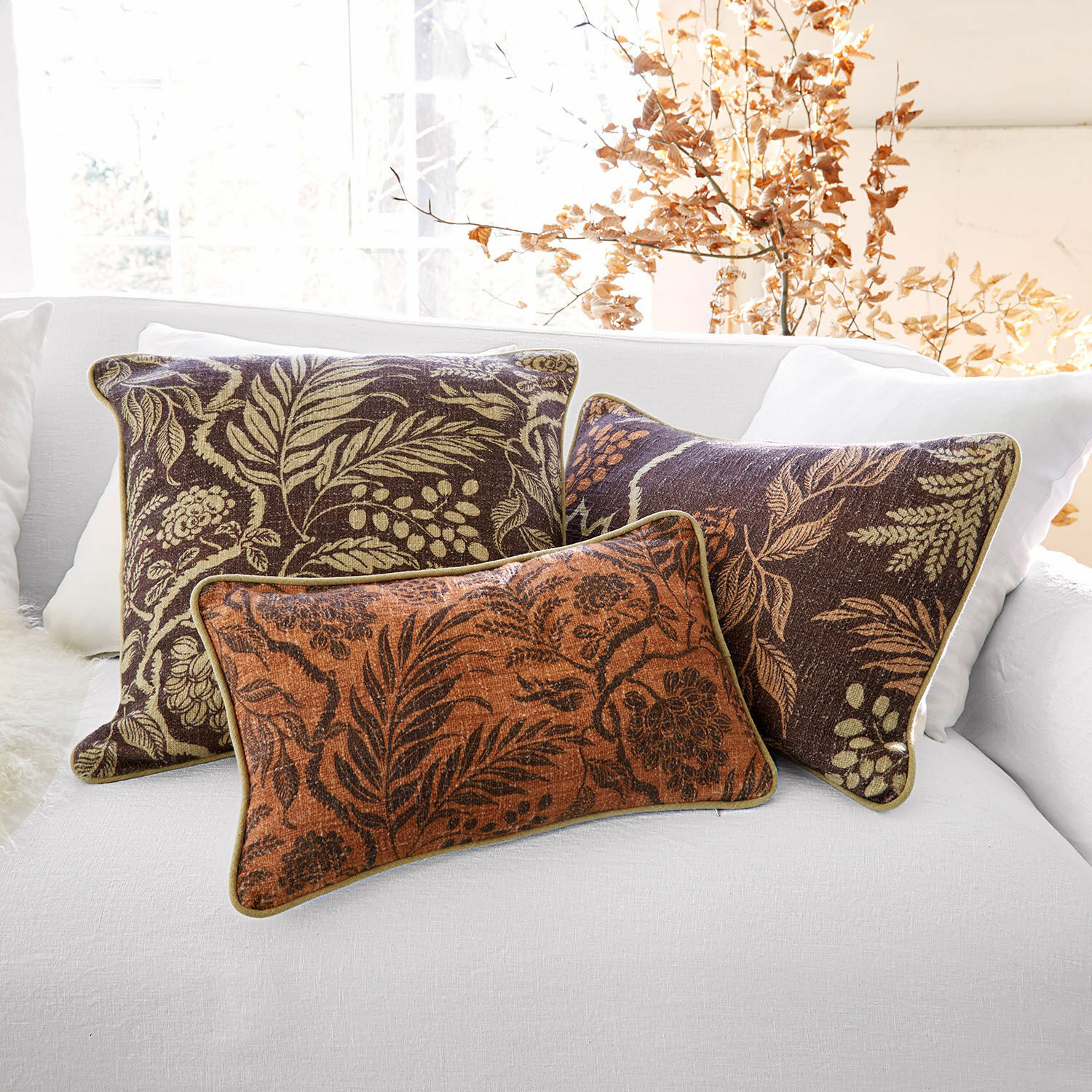 Set di 3 copricuscini Carivelle Set di tre cuscini decorativi con motivo foglie in tonalità marroni e arancioni su divano bianco