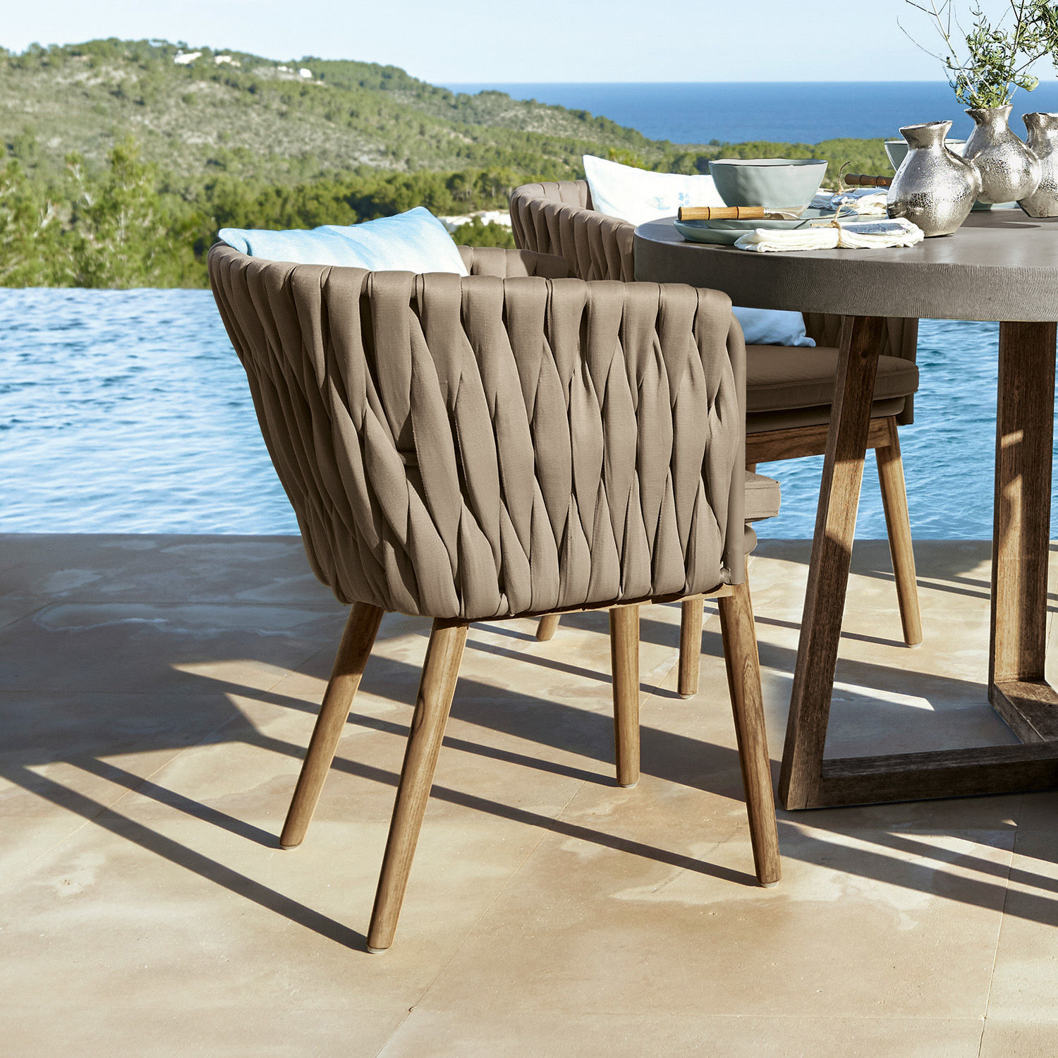 Sedia Uchon Sedia da esterno beige con schienale intrecciato e gambe in legno su terrazza con vista mare