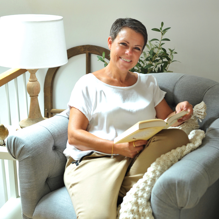 Donna con maglietta bianca e pantaloni beige legge un libro su un divano grigio comodo in una stanza luminosa