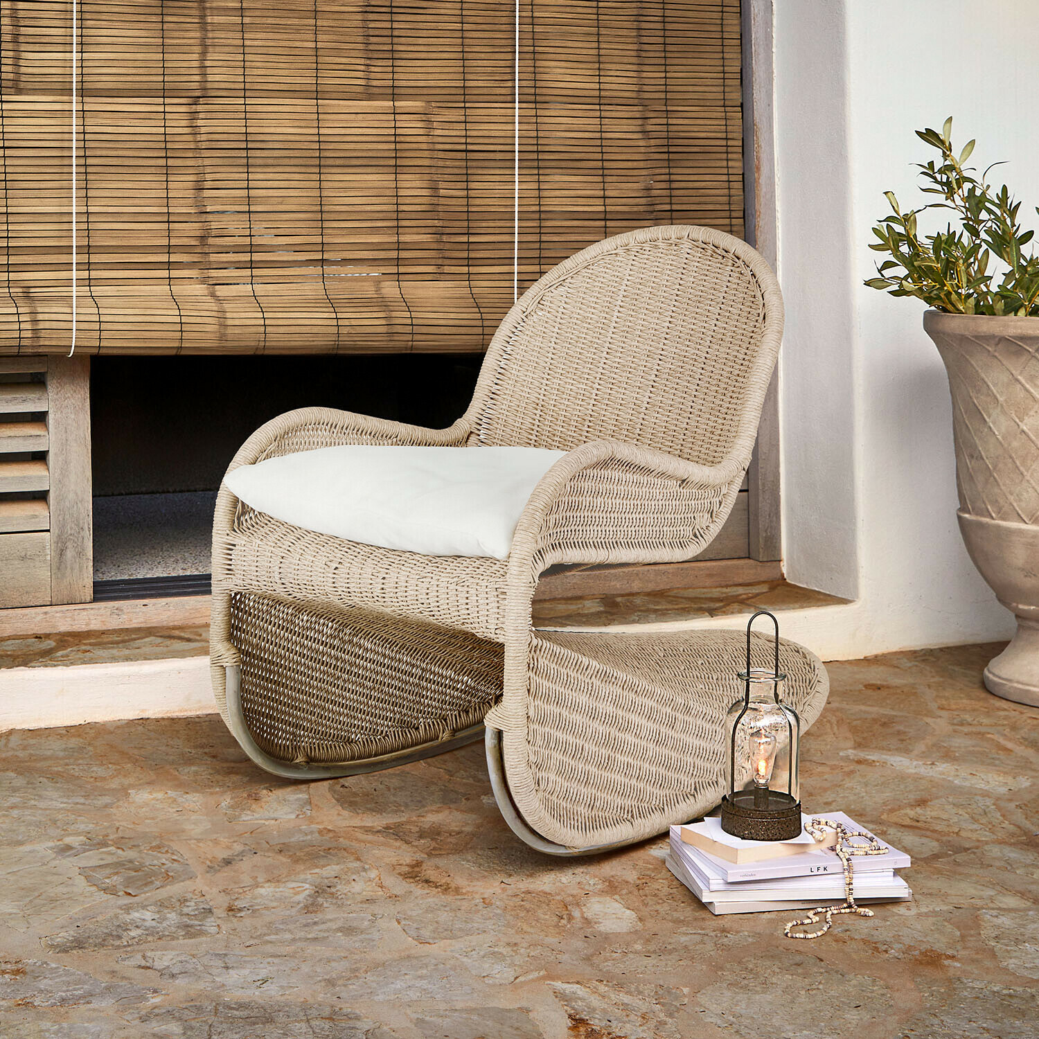Poltrona in rattan beige con cuscino bianco su pavimento in pietra davanti a tenda in bambù