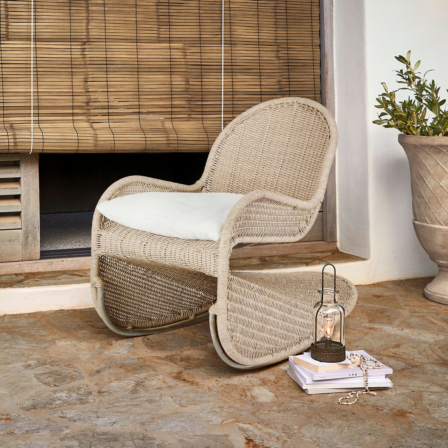 Sedia a dondolo Fernleigha Poltrona in rattan beige con cuscino bianco su pavimento in pietra davanti a tenda in bambù