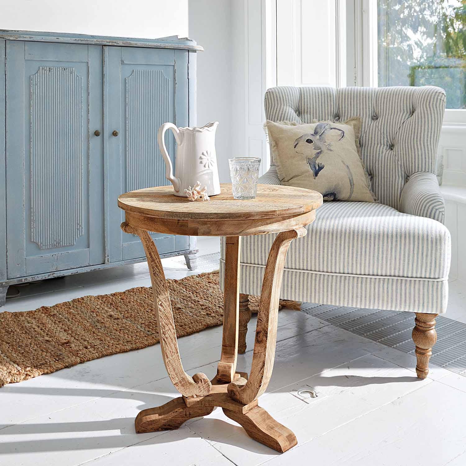Tavolino rotondo in legno di mango naturale in soggiorno luminoso con poltrona a righe e credenza azzurra