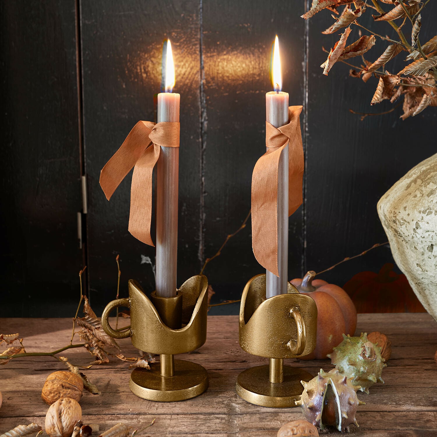 Due candelabri in metallo dorato con candele grigie e nastri arancioni su tavolo in legno rustico
