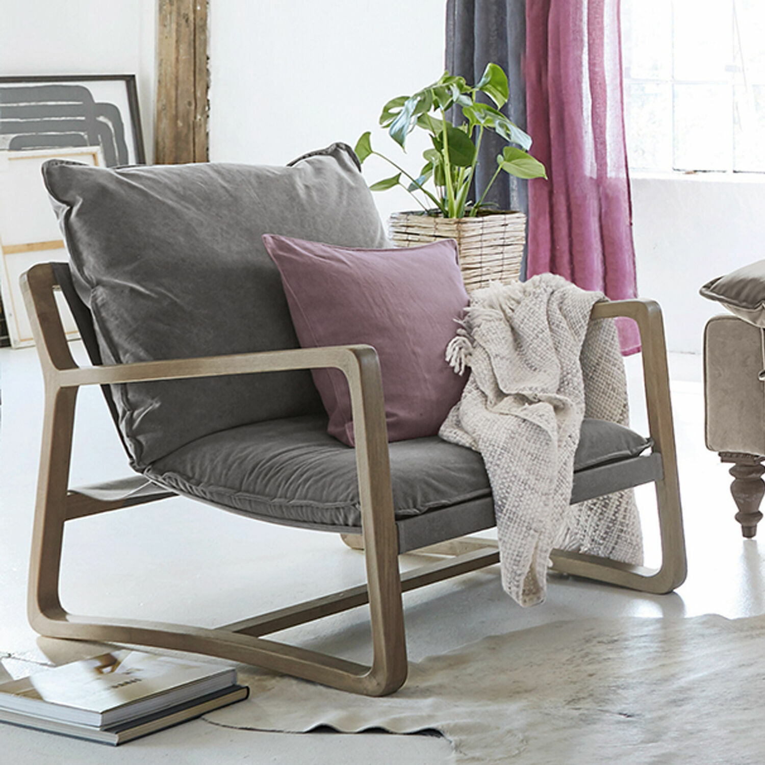 Poltrona Jowna Poltrona con rivestimento grigio e struttura in legno, cuscino viola e coperta beige in soggiorno