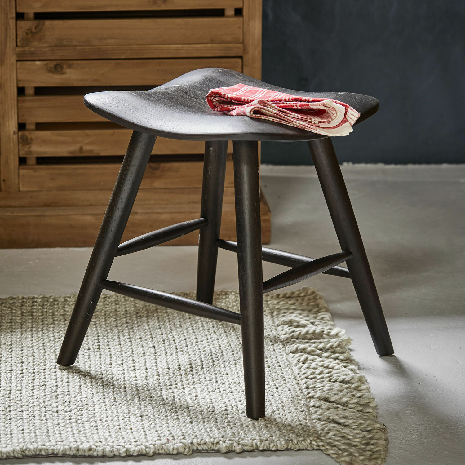 Sgabello Voiláren Sgabello in legno nero con seduta curva su tappeto chiaro, tovagliolo decorativo rosso piegato