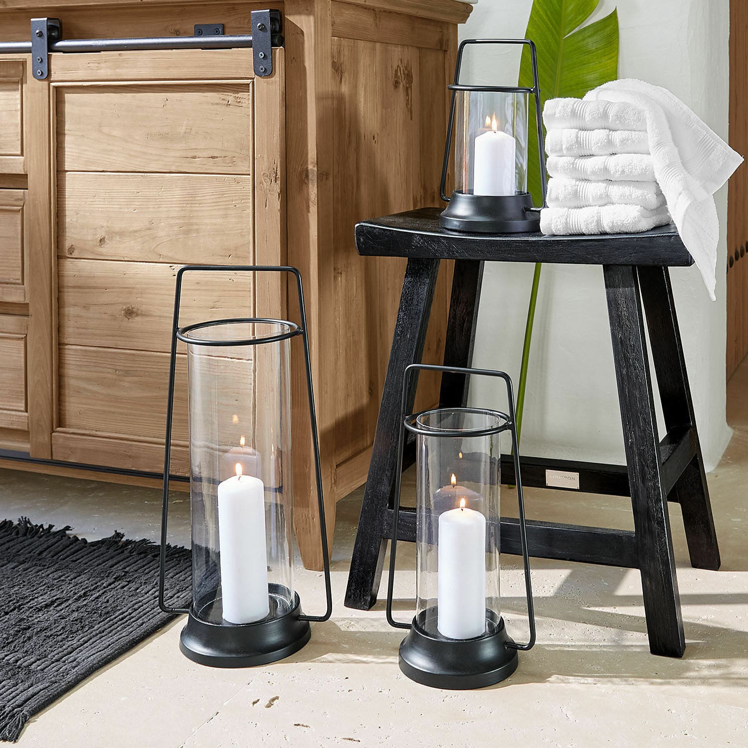 Set di 3 lanterne Mirandel Set di tre lanterne in metallo nero con candele bianche e asciugamani su uno sgabello nero