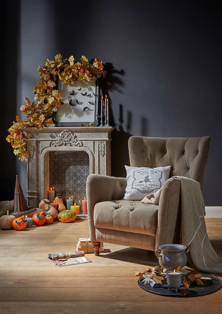 Poltrona beige imbottita con cuscino e coperta accanto a camino decorato con zucche e decorazioni autunnali