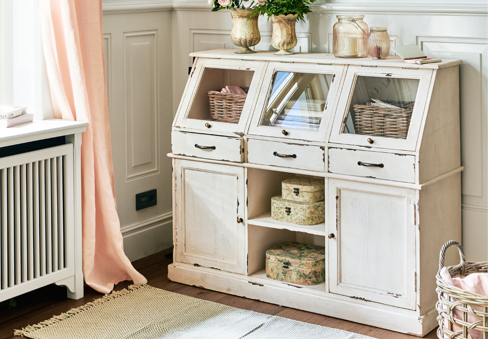 Credenza in legno bianco stile shabby chic con cassetti in vetro e decorazioni in una stanza luminosa.
