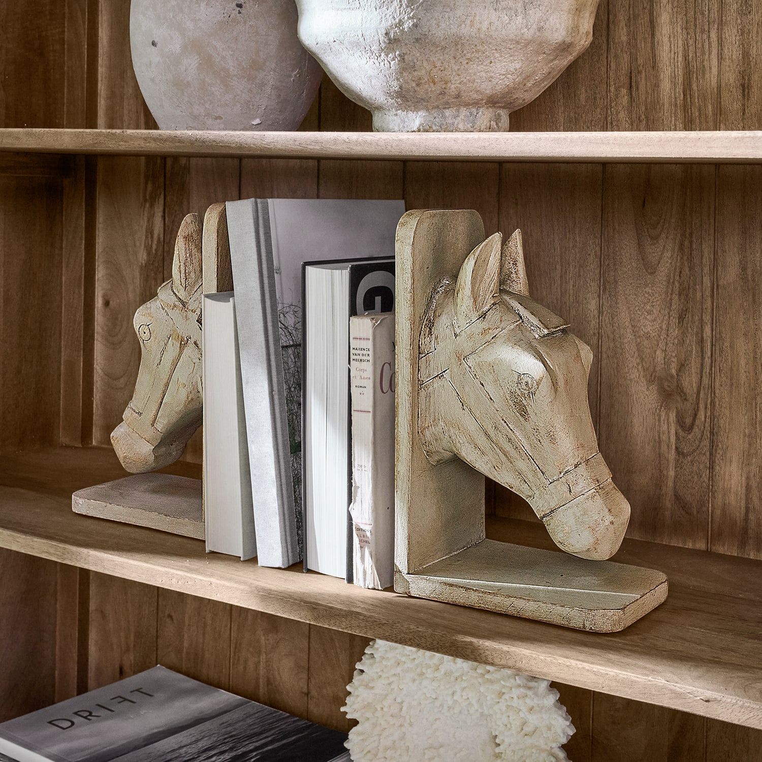 Coppia di fermalibri in legno beige a forma di testa di cavallo su mensola in legno con libri