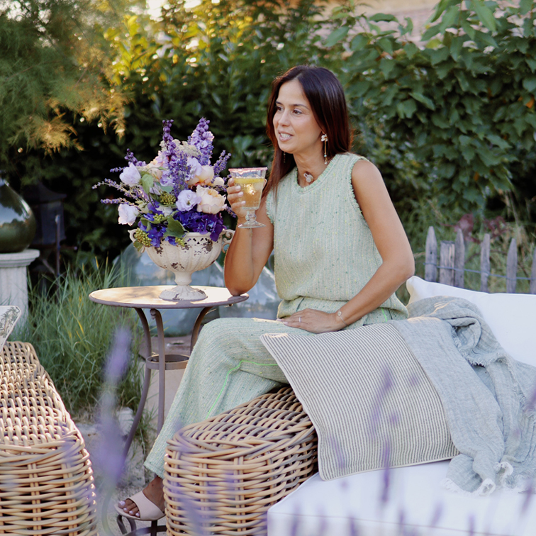 Donna con vestito verde seduta su mobile in vimini in giardino con bouquet di fiori e bicchiere di vino