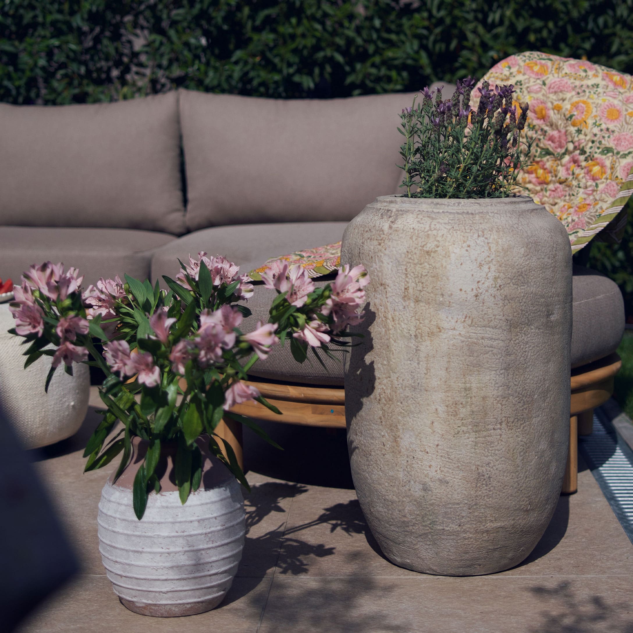 Vaso grande e piccolo in pietra chiara con fiori su terrazza con divano grigio e coperta colorata