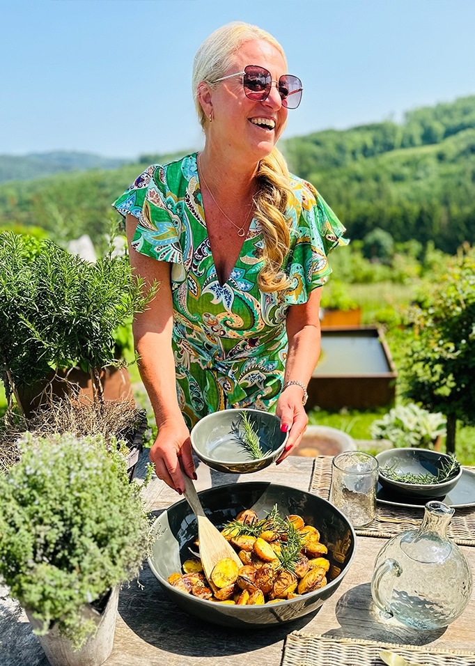 Donna in abito verde che serve patate al forno in ciotola di ceramica all'aperto con erbe e natura