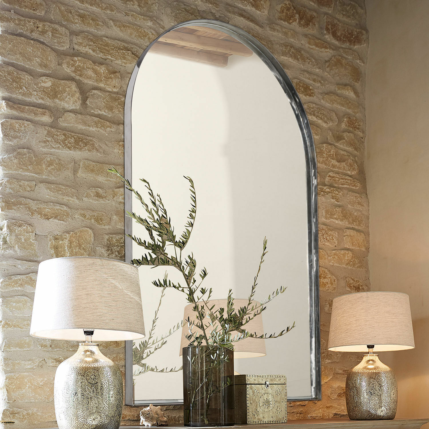 Specchio Kellin Grande specchio con cornice in metallo su muro di pietra con lampade e decorazioni su mensola in legno