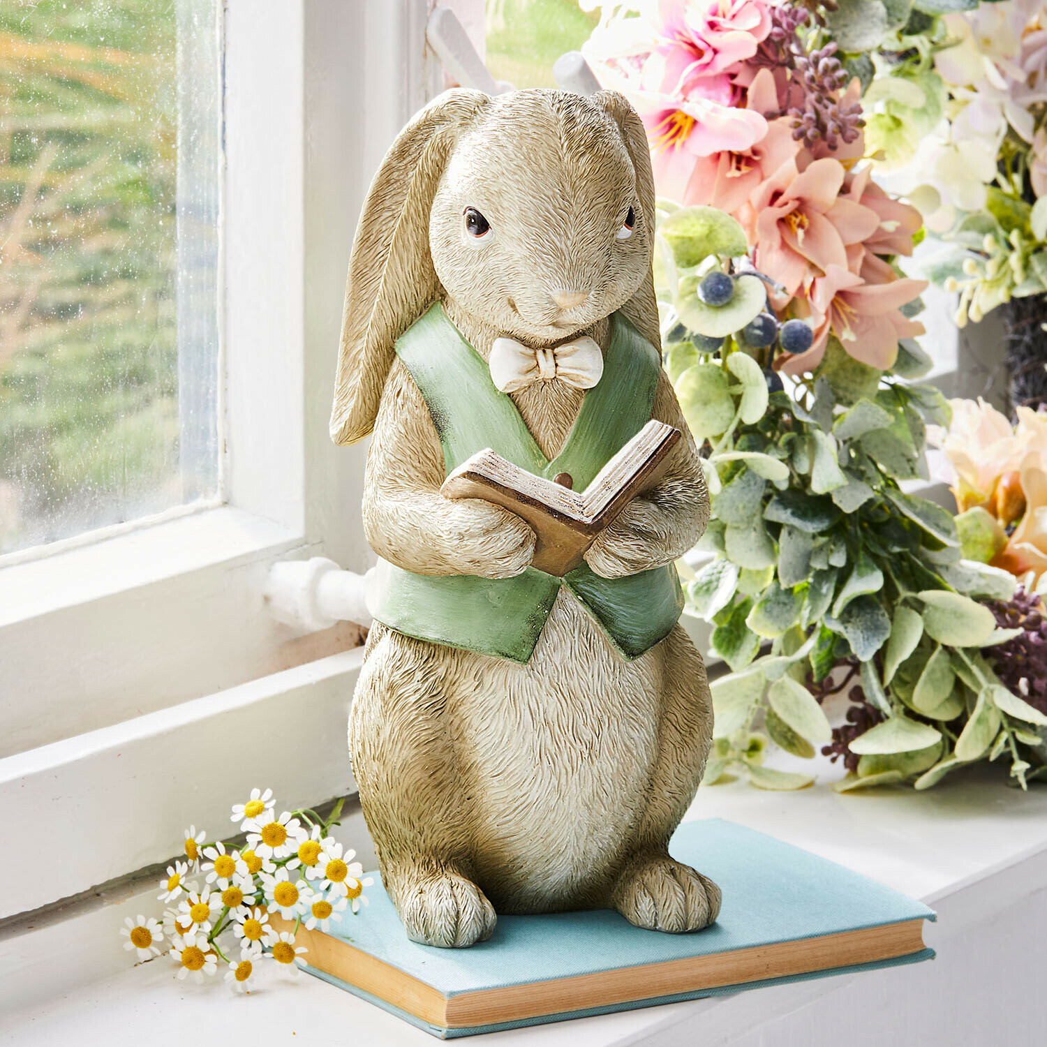 Statua di coniglio in resina con gilet verde che legge un libro su davanzale con fiori