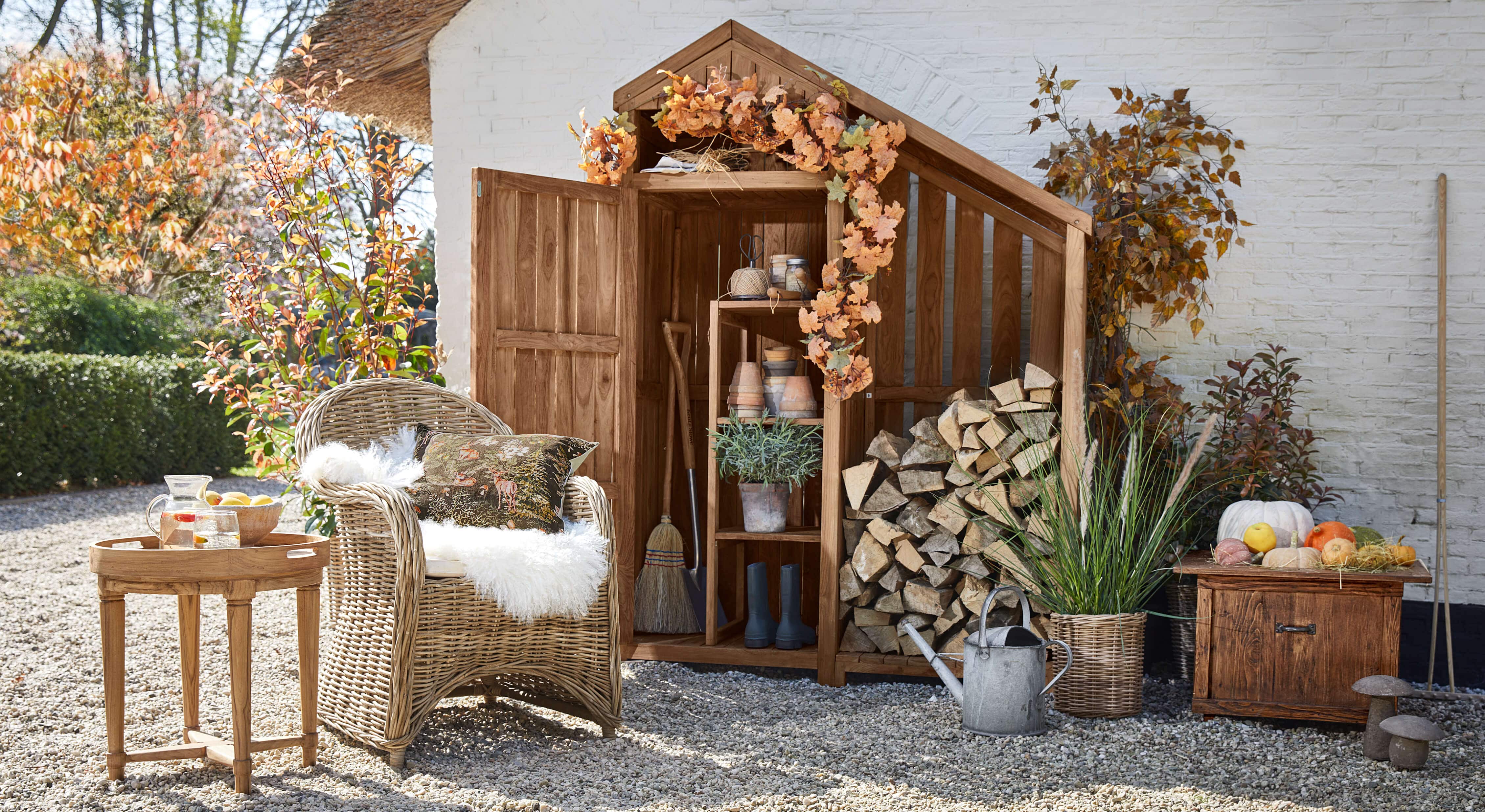 Casetta da giardino in legno con decorazioni autunnali, poltrona in vimini e tavolino con brocca su ghiaia
