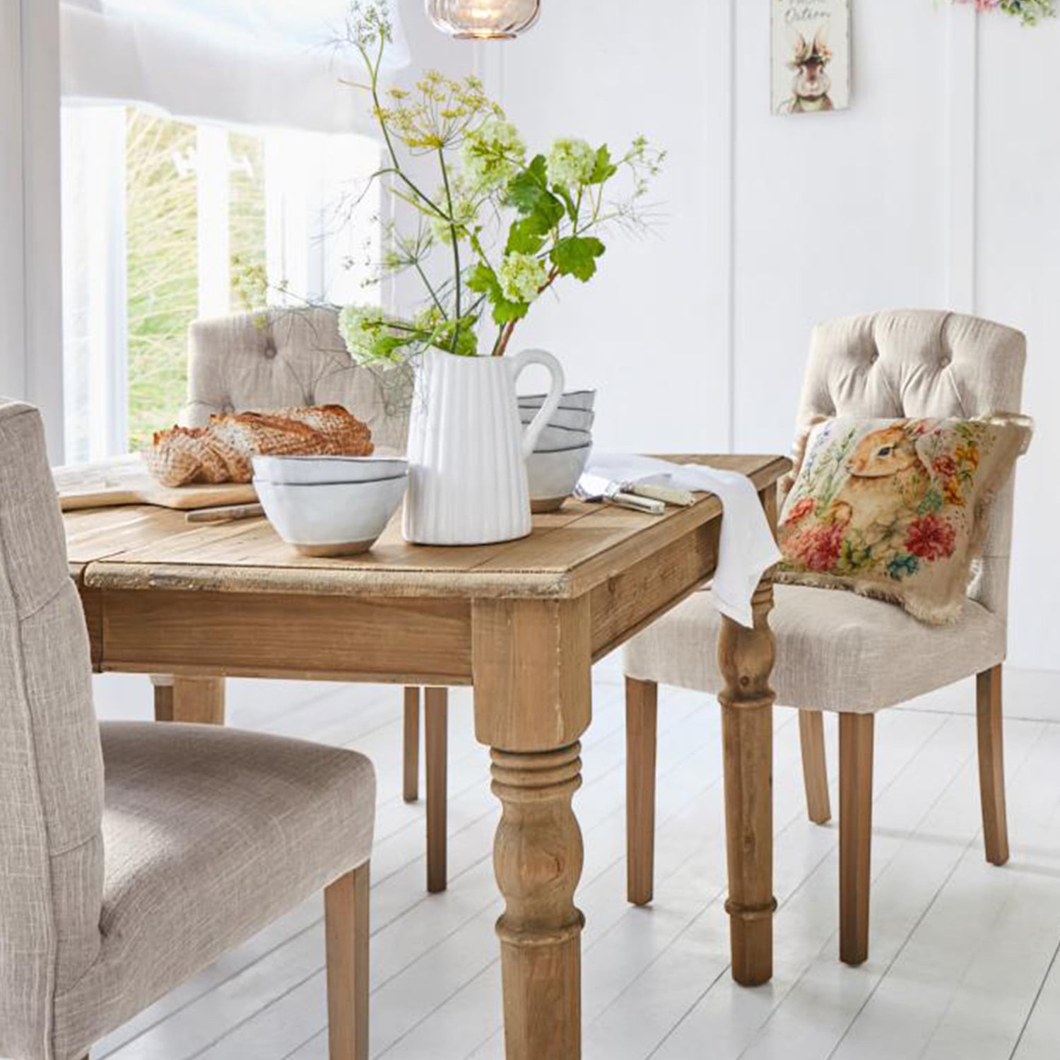 Tavolo in legno chiaro con brocca decorativa e ciotole, sedie imbottite beige in sala da pranzo luminosa