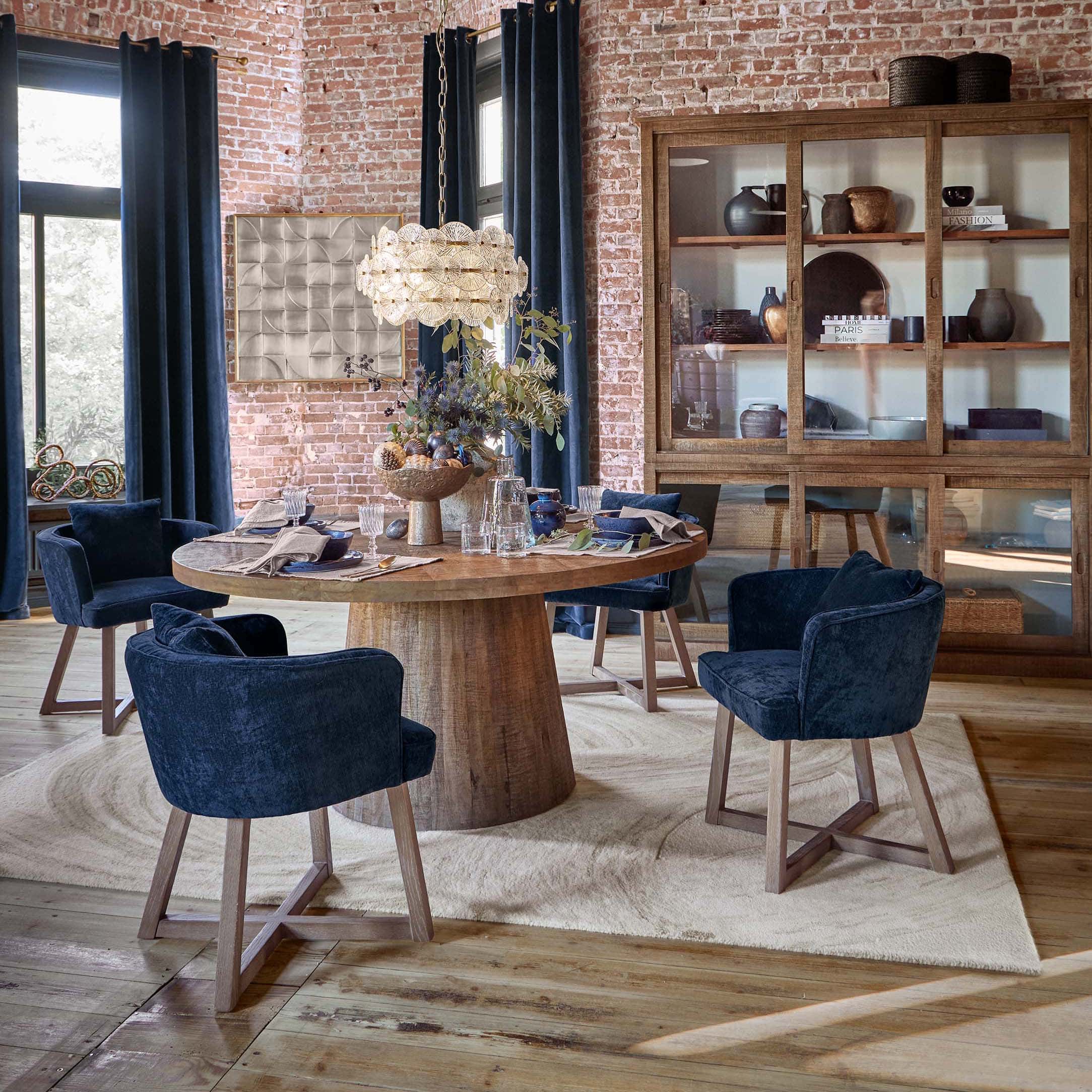 Sala da pranzo con tavolo rotondo in legno e sedie in velluto blu scuro davanti a muro in mattoni e vetrina in legno