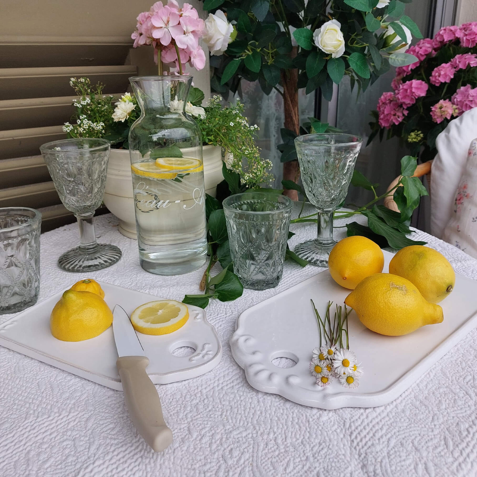 Caraffa di vetro con acqua al limone su tovaglia bianca, bicchieri decorativi e taglieri in ceramica bianca