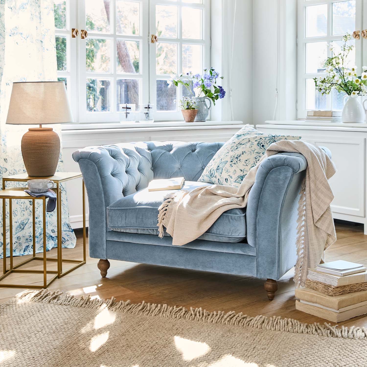 Divano due posti in velluto blu con cuscino floreale e coperta beige in soggiorno luminoso con pavimento in legno