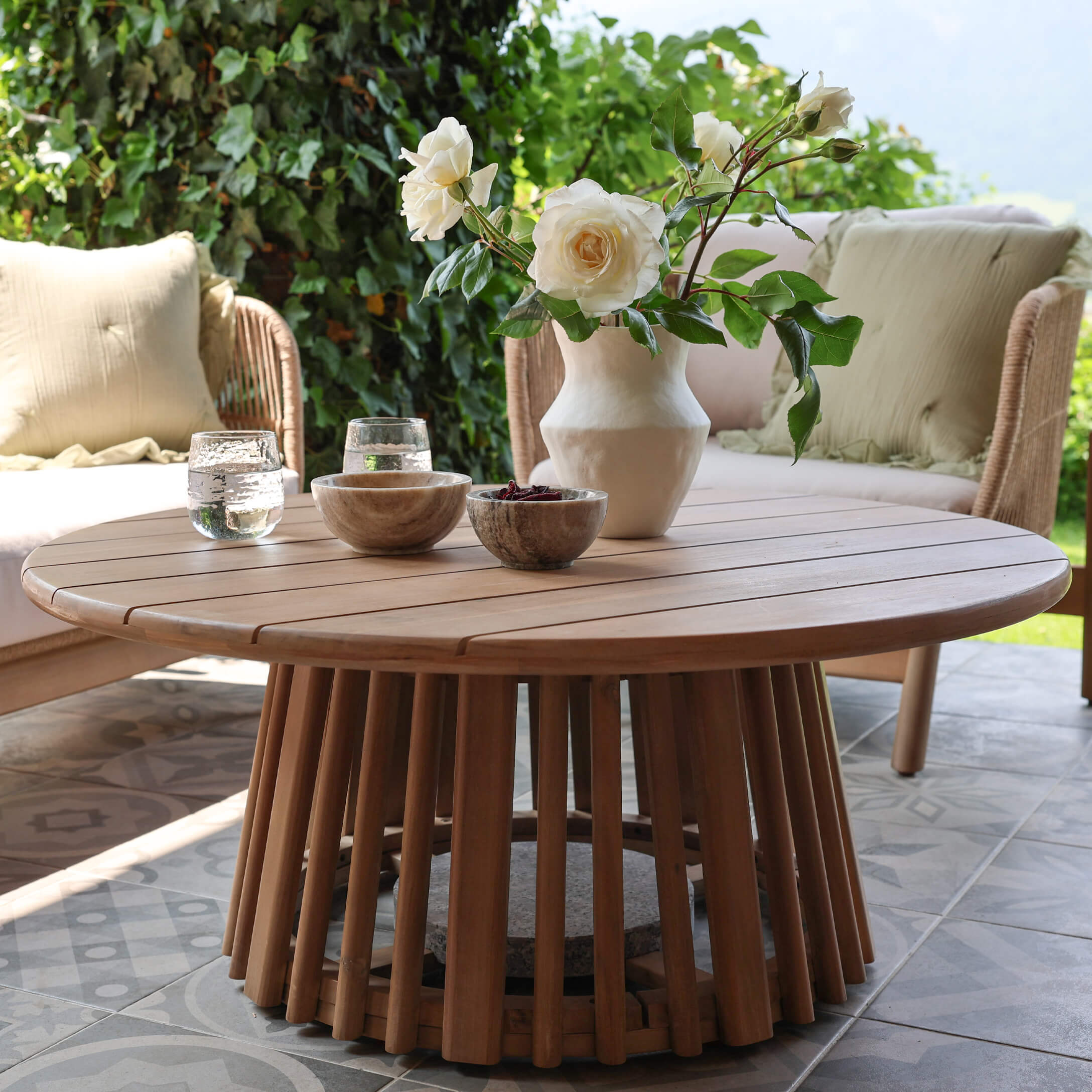Tavolino rotondo in legno di teak naturale su terrazza con decorazione floreale e ciotole