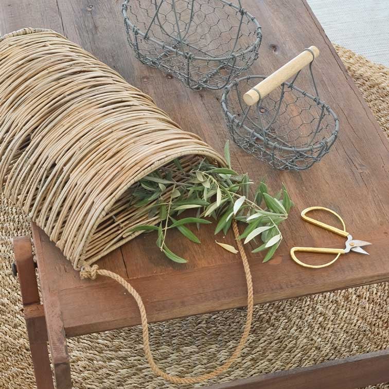Cesto in vimini con manico in corda su tavolo di legno con rami verdi e forbici da giardino dorate