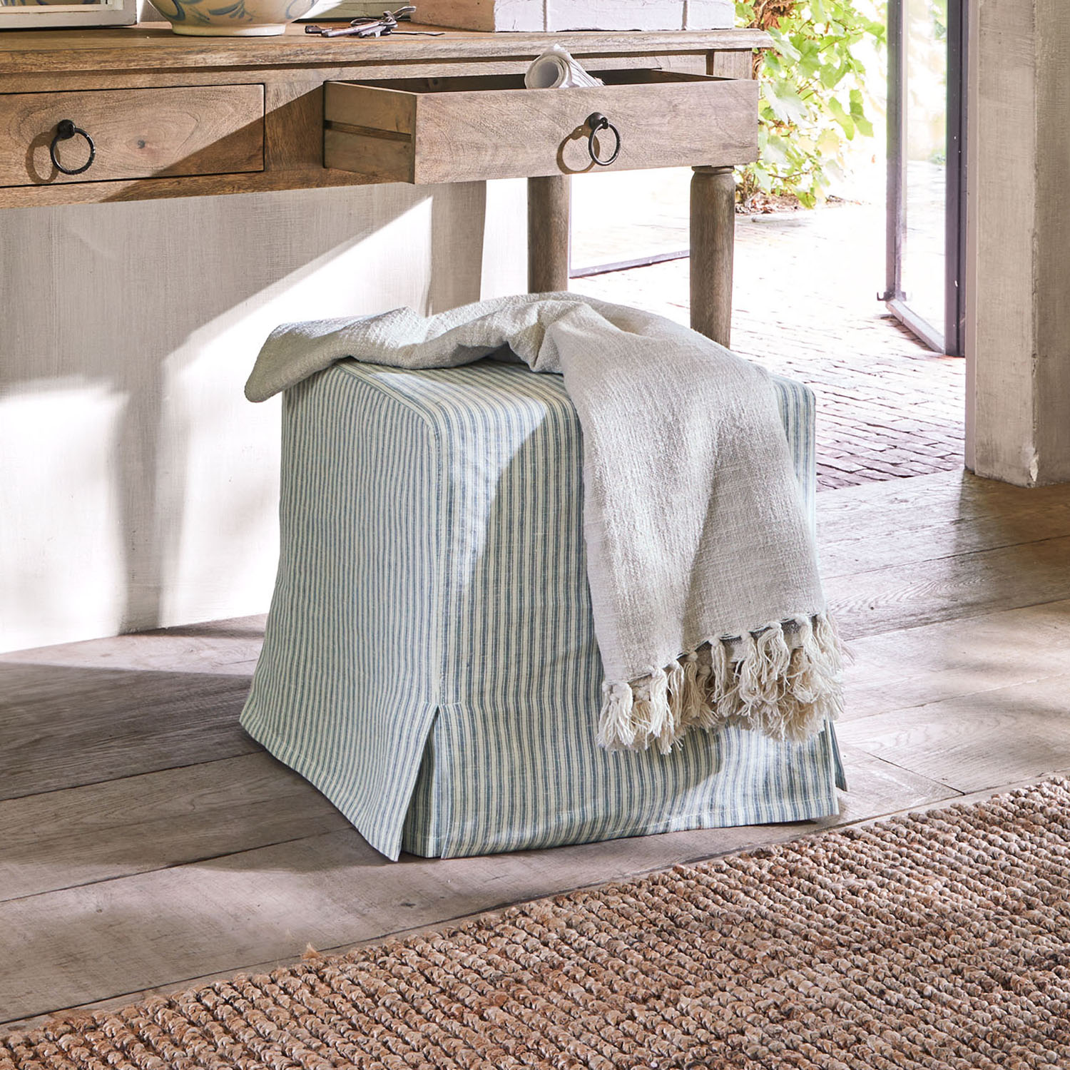 Pouf quadrato in cotone a righe blu e bianco con coperta chiara con frange su pavimento in legno