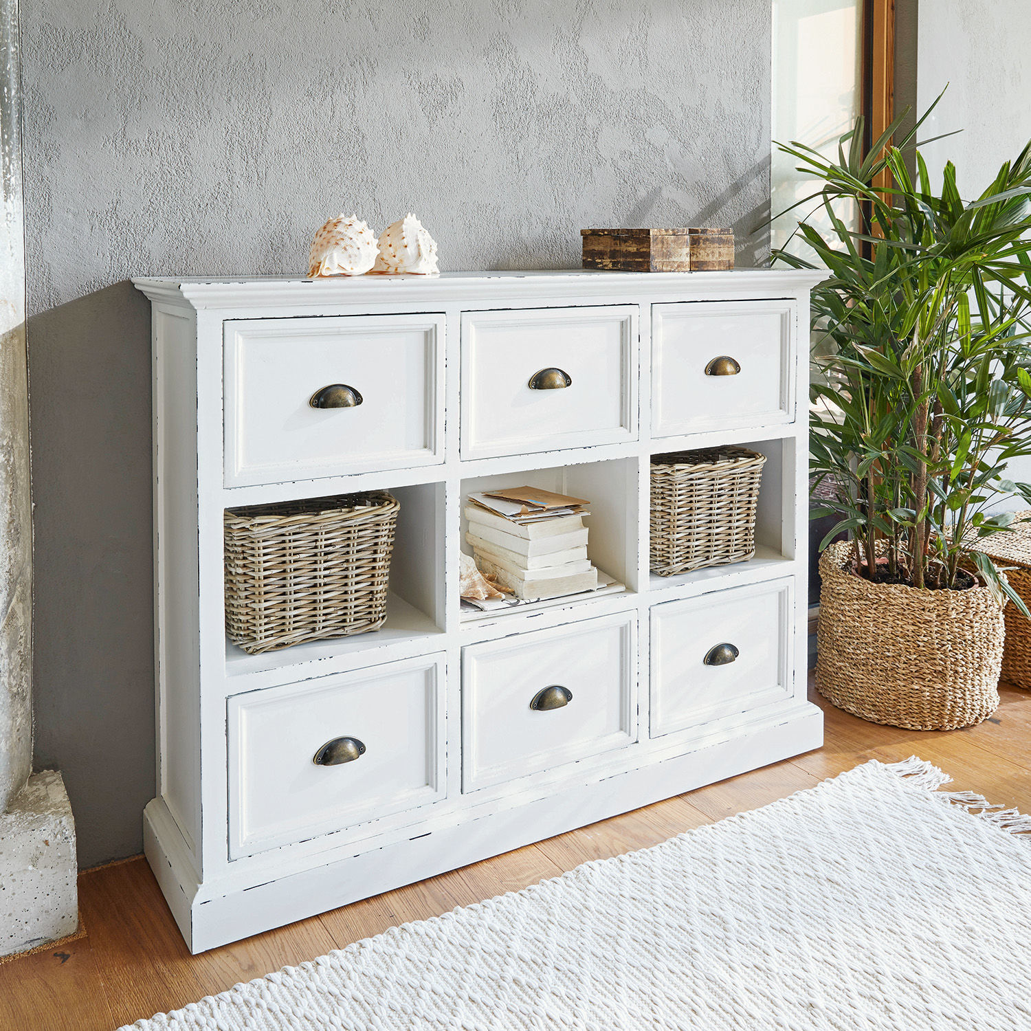 Mobile con cassetti Remilly Comò bianco in legno con sei cassetti e tre ripiani aperti, decorazioni e pianta accanto