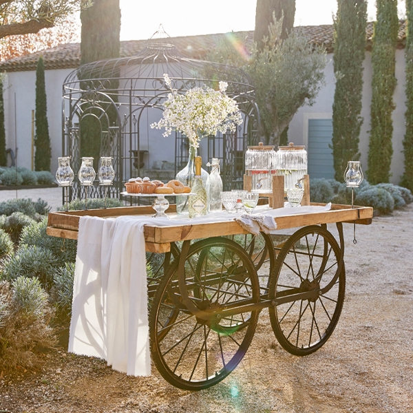 Carrello in legno con ruote in metallo, tovaglia bianca e fiori decorativi in giardino soleggiato
