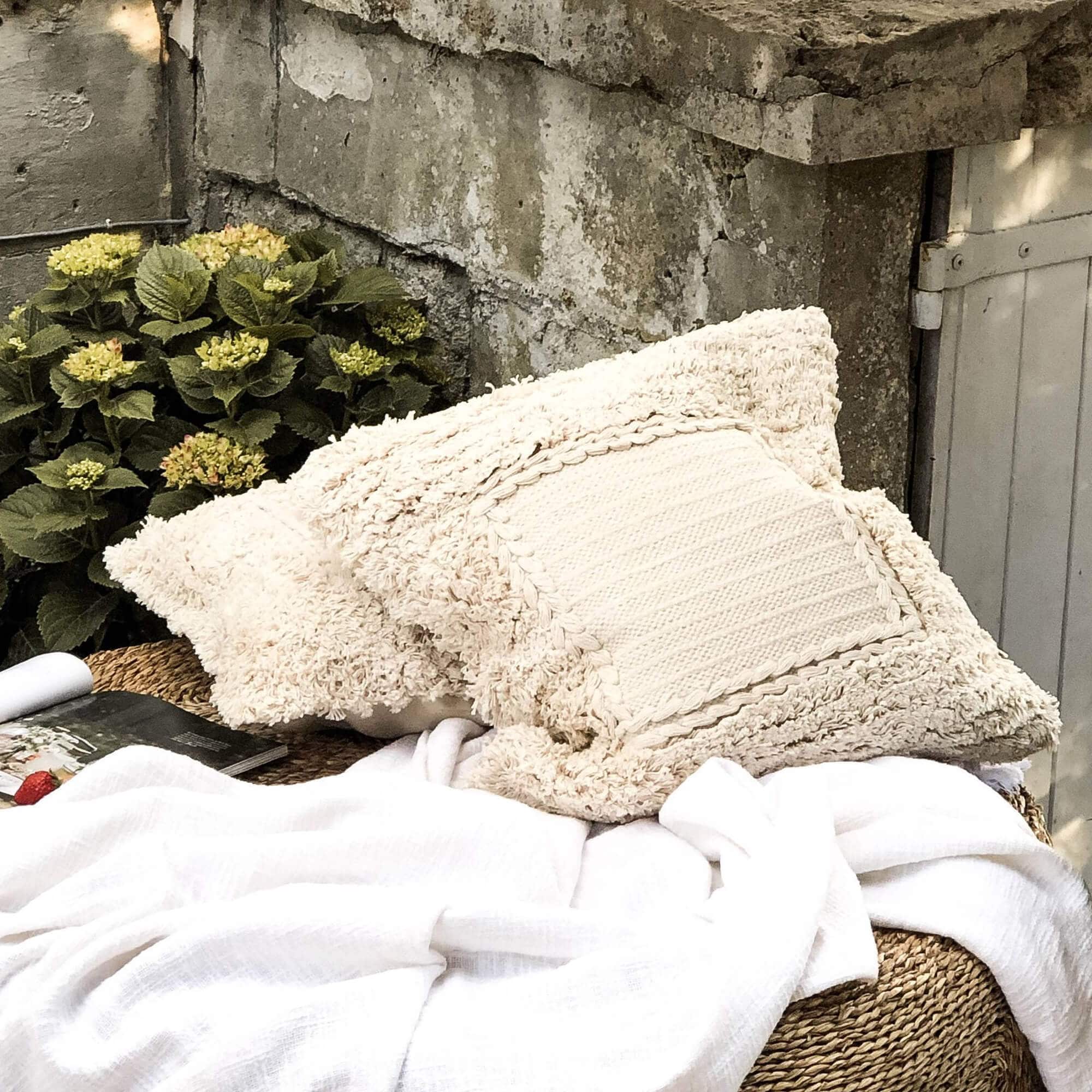 Cuscino decorativo beige in materiale morbido su mobile intrecciato con coperta bianca