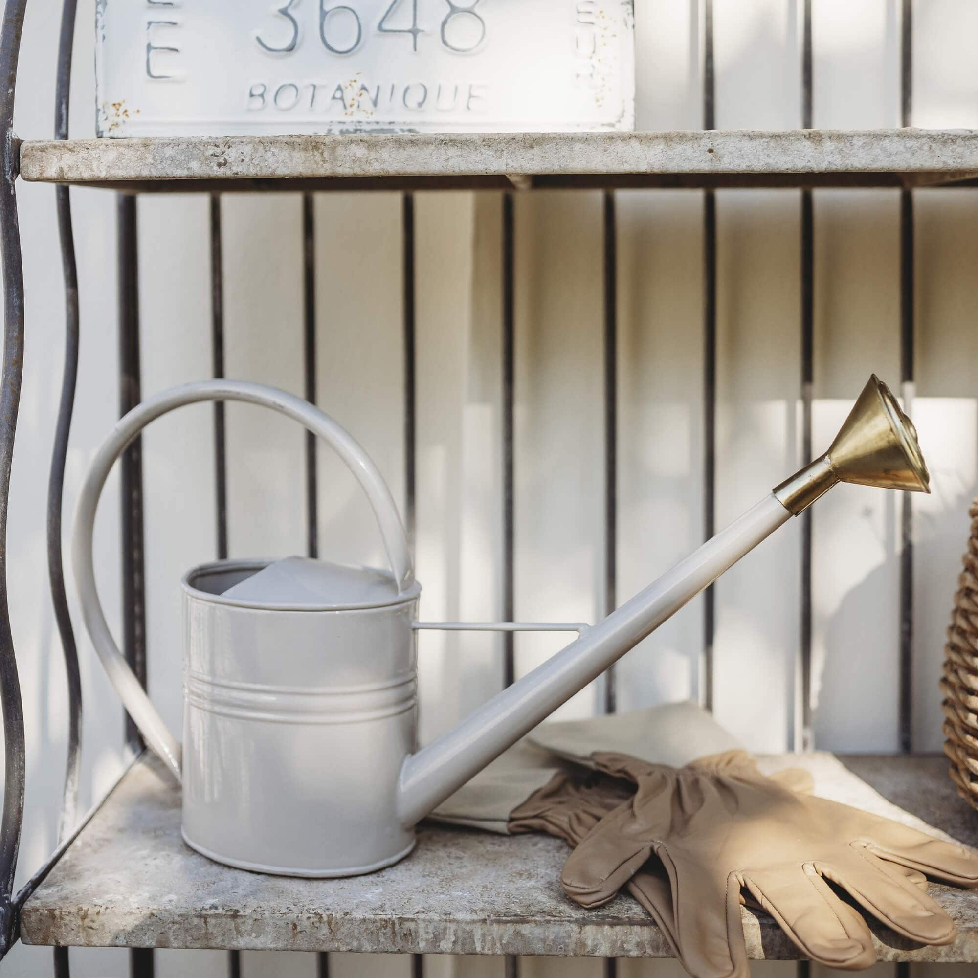 Annaffiatoio in metallo beige con beccuccio in ottone e guanti da giardinaggio su mensola in pietra