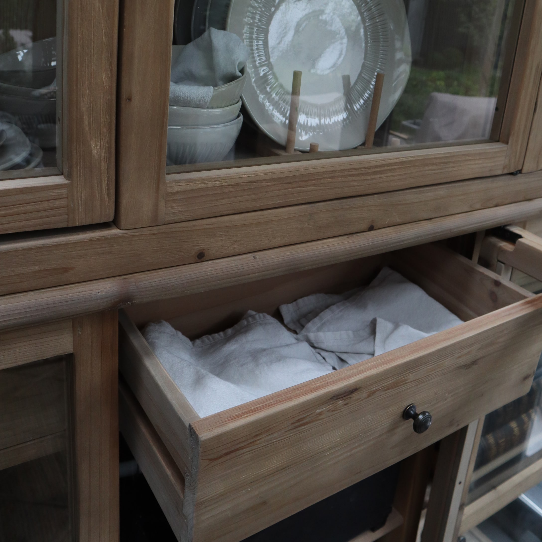 Mobile in legno con cassetto aperto e tessuti bianchi, ceramica bianca dietro porta in vetro