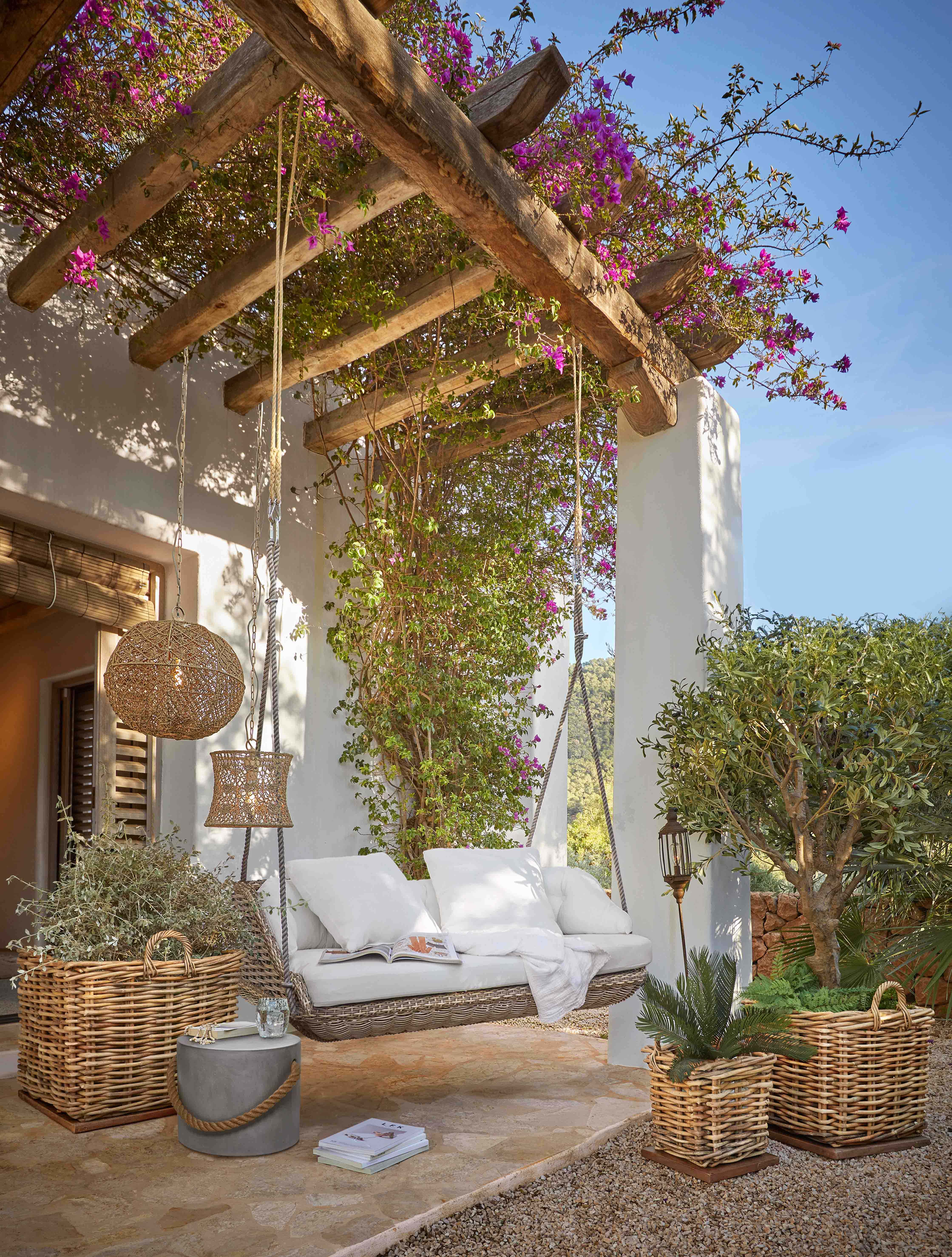 Dondolo in rattan con cuscini bianchi sotto una pergola in legno con fiori rosa in fiore