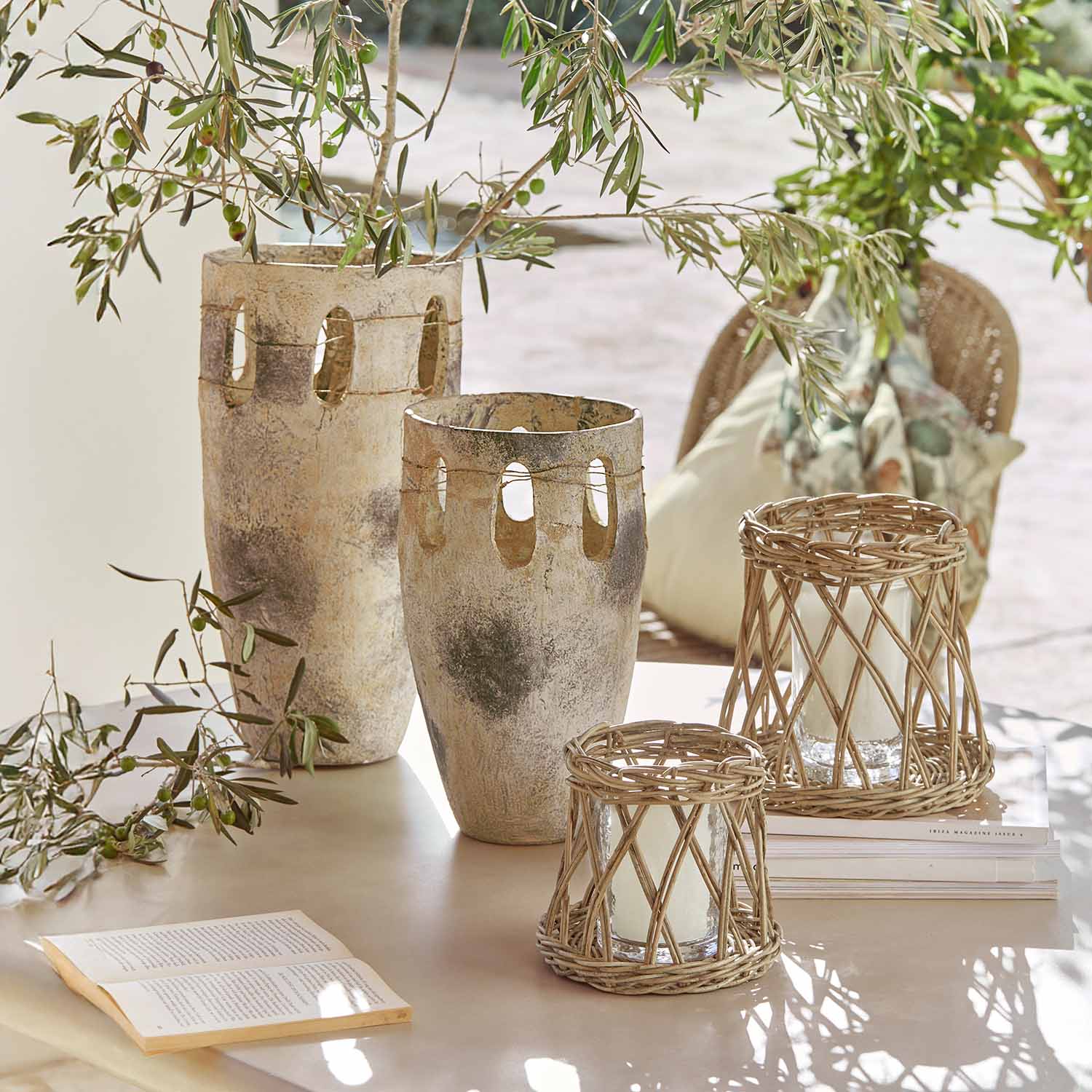 Due grandi vasi in ceramica e due lanterne in rattan su un tavolo con rami verdi e un libro aperto