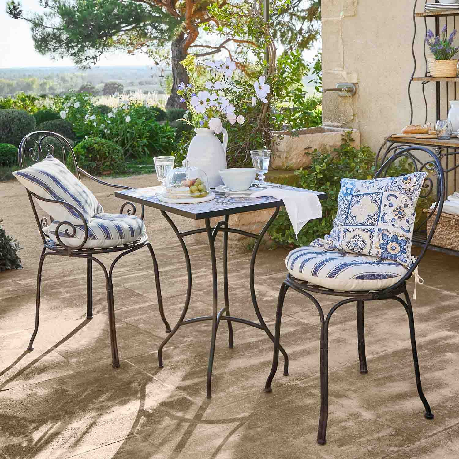 Tavolo quadrato in metallo con due sedie con cuscini blu su terrazza soleggiata in giardino