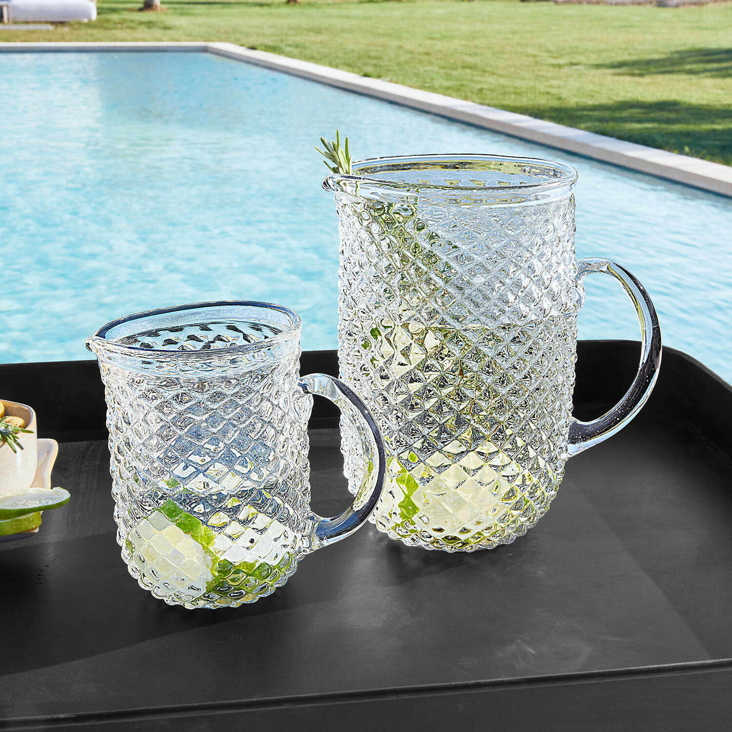 Coppia di caraffe Ivorias Caraffa e bicchiere in vetro trasparente con motivo a diamante, con fette di lime, su vassoio nero vicino alla piscina