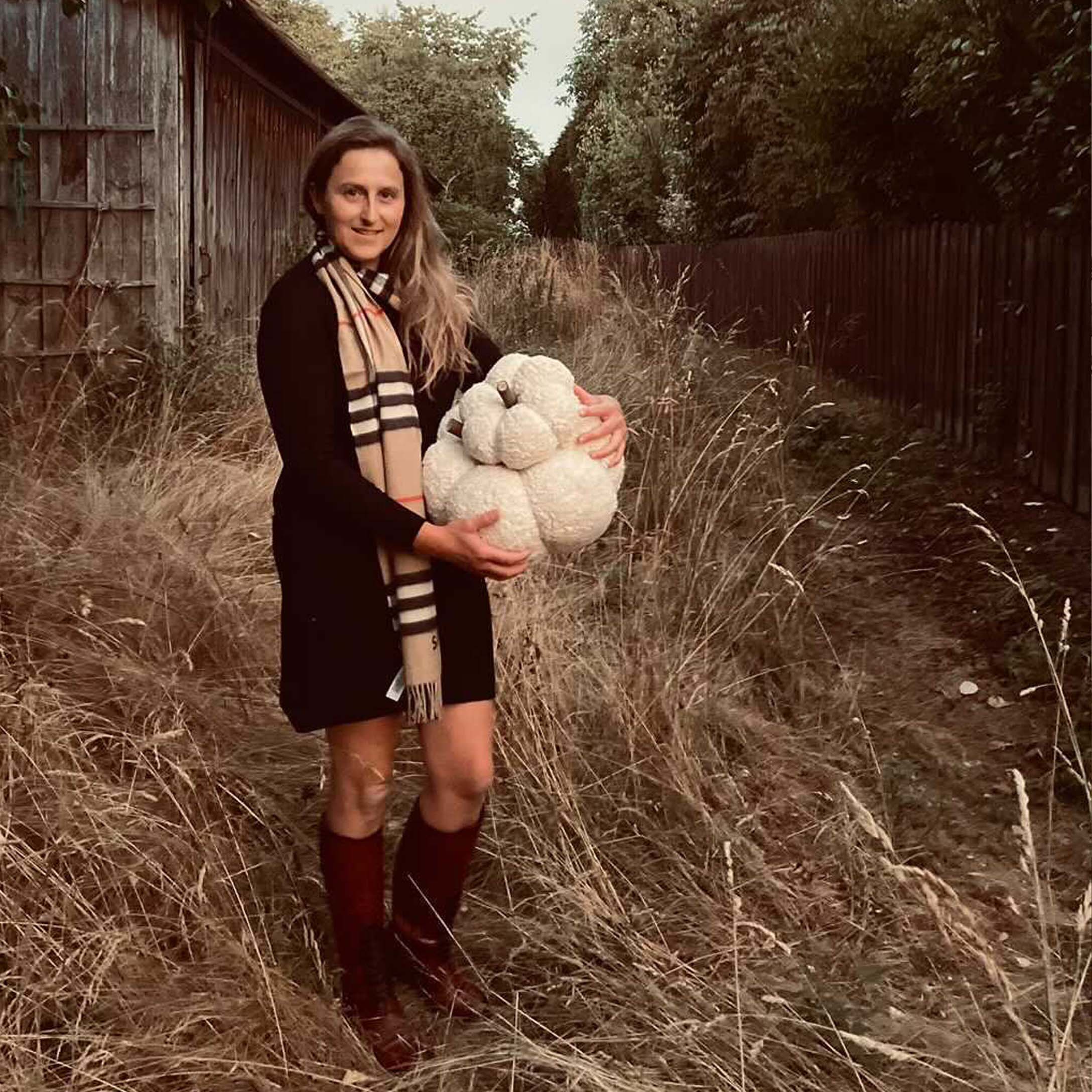 Donna con vestito nero e sciarpa che tiene una zucca decorativa bianca in un campo autunnale
