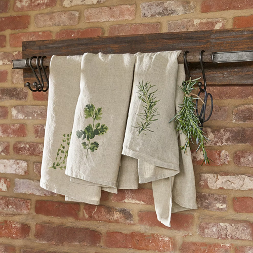 Strofinacci da cucina in lino colore naturale con stampa di erbe appesi a supporto in legno su muro di mattoni
